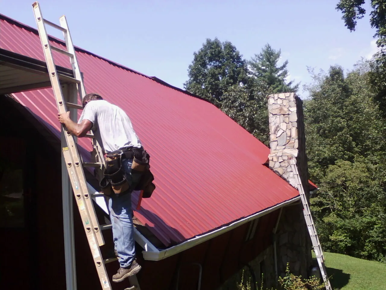 Roofer on a ladder beside a red metal roof and stone chimney — Soffit & Fascia Repair by Roofing Brothers in Ashford